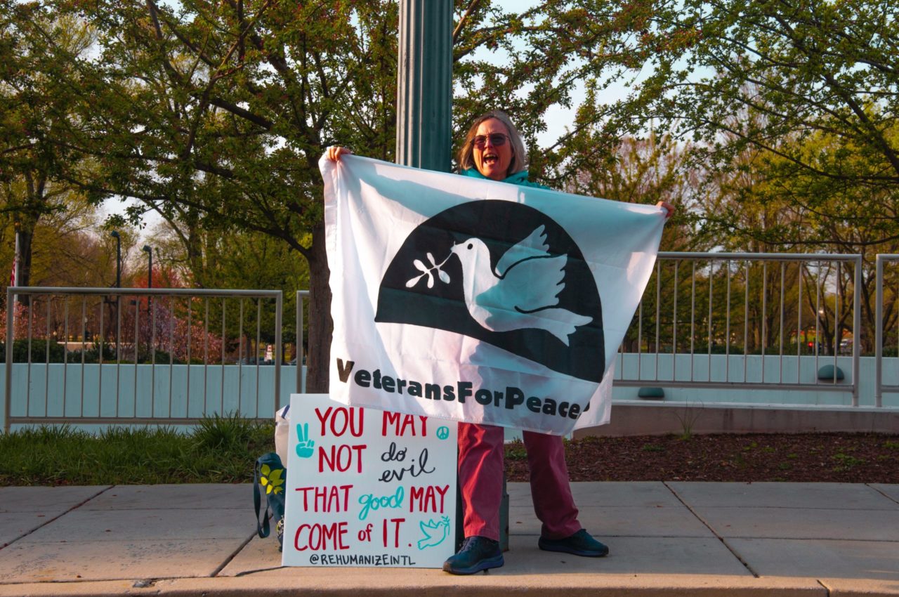 Person stands holding flag that reads Veterans for peace with dove in the middle.