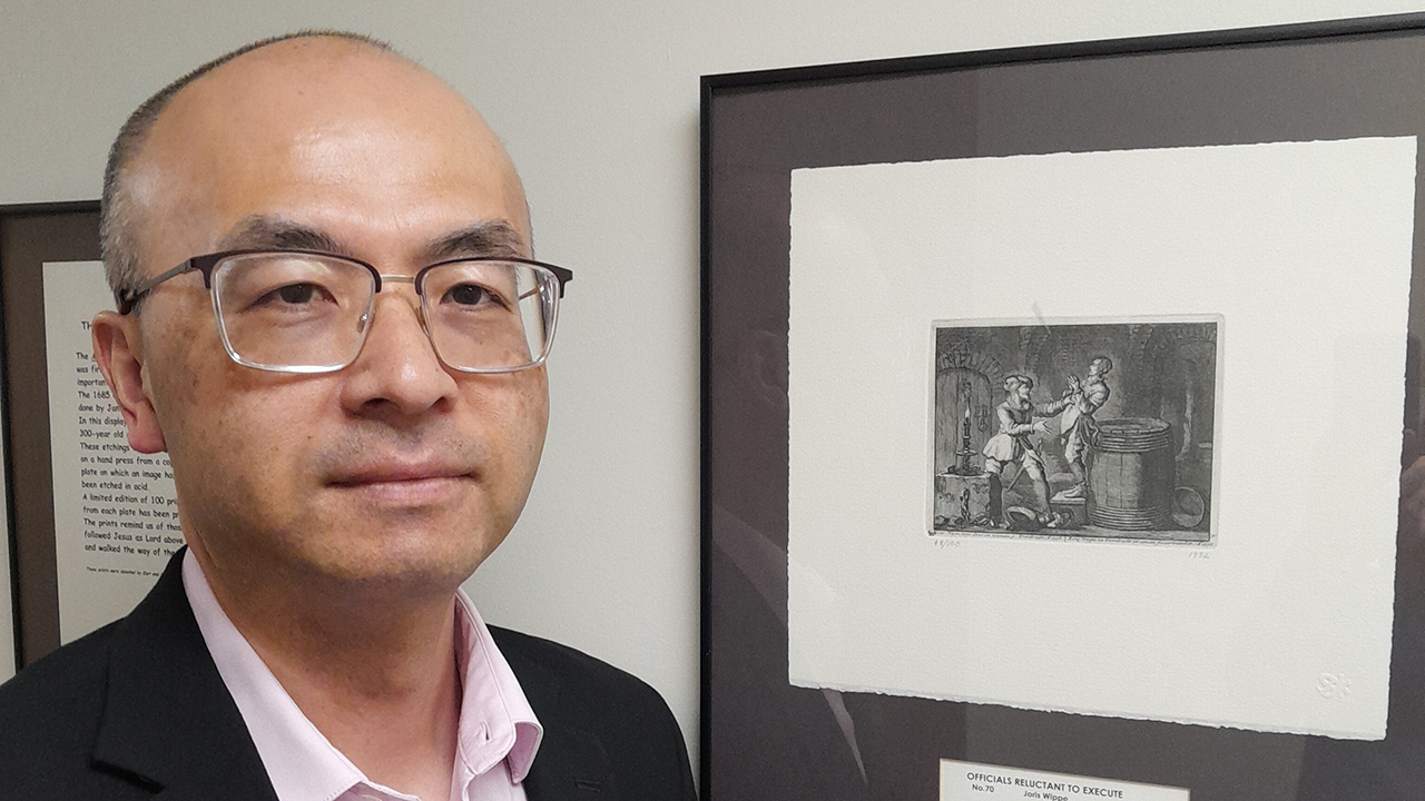 Shuji Moriichi with one of the framed prints of Martyrs Mirror art on a wall at First Mennonite Church of Iowa City, Iowa. — Courtesy of Shuji Moriichi